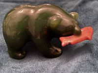 Side view of small dark green jade bear holding a pink jade salmon in its mouth on a dark fabric background.
