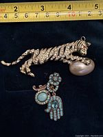 Full view of rhinestone leopard brooch holding oval pearl-like bead with copper turquoise hamsa charm below it on black background with measuring tape