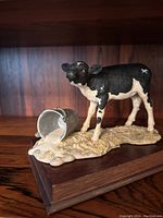 View of the small calf sculpture showing detailed coloring, tipped milk churn, textured base and dark wooden platform.