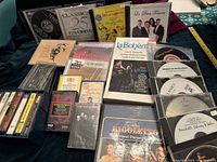 Photo shows a collection of CDs and cassette tapes with opera and classical music titles such as La Bohème, Rigoletto, and Royal Philharmonic Orchestra. Various jewel cases and cardboard sleeves are visible.