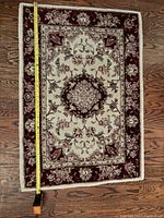 Full view of one Persian wool rug showing measuring tape indicating length of approximately 3 feet on a dark wood floor, rug with cream center and burgundy floral border pattern