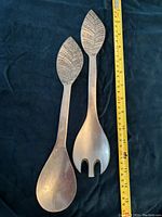Two pewter salad servers, one spoon and one fork, each with a leaf motif handle, lying next to a measuring tape showing length around 10 inches.