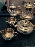 Close-up shot showing the silver-plated coffee pot, sugar bowl, creamer, and part of the teapot, highlighting the ornate lids and handles with scalloped and floral detailing.