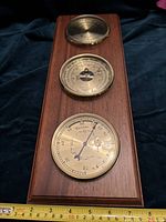 Full view of the wooden wall-mounted instrument panel showing three brass-cased round instruments: hygrometer, barometer, and thermometer, with a measuring tape showing about 15 inches in height.