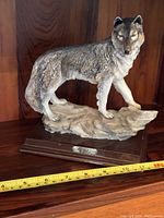 Side angle photo showing the full wolf sculpture standing on sculpted ground, mounted on a wooden base with a nameplate labeled 'Wolf'. Measurement tape shows the sculpture length approximately 10 inches.