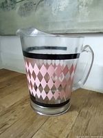 Clear glass water jug with pink and black diamond pattern, two black bands, handle and spout, angled view on wooden surface