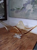 Side view of signed Chalet amber colored art glass bowl with wavy edges and clear sculpted base on wooden surface