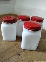 Photo of four vintage white glass spice jars with red metal lids on wood surface. One lid has holes for sprinkling.