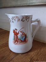 Side view of white china jug with figurative decoration of two children, showing ornate gold scrollwork near the rim and curved handle.