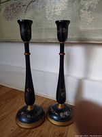 Pair of vintage Danish wooden candlesticks standing upright on a wooden floor against a white wall. Both show the hand-painted floral design and gold accents on dark wood.