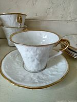 Close-up image of single white porcelain teacup and saucer with dimensional oak leaf embossing and gold trim on rim and handle.