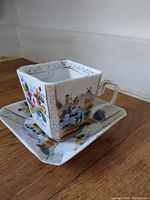 Square handpainted teacup and saucer on wooden surface showing scenic and floral artwork with gold accents.