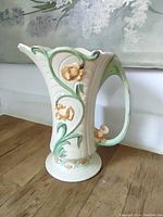 Photo of off-white ceramic vase with raised yellow daffodils and green leaves, flared rim and curved handle sitting on wooden surface.