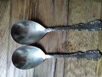 Two antique silver spoons laid side by side showing full length and detailed handle design.