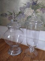 Photo showing two large clear glass apothecary jars with lids on a wooden surface with a floral background.
