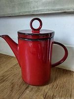 Red ceramic coffee pot with lid and circular handle on top, placed on a wooden floor near a wall.
