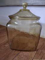 Vintage amber glass canister with lid, showing embossed floral patterns on jar body and lid, placed on wooden floor.