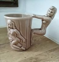 Light tan ceramic mug with nude female figure handle and raised nude figure relief on mug sides, shown on wood surface against white wall.