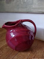 Photo showing glossy red ceramic jug with rounded body, prominent handle, wide pouring spout, sitting on a wooden surface against a wall.