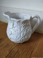 White ceramic jug shaped and textured like a cabbage leaf, showing the handle, spout, and embossed leaf detail.