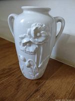 Front view of white ceramic vase showing detailed raised poppy flower design with two handles.