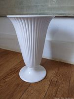 Front and side angled view of tall white ceramic vase with vertical fluting on wooden floor.