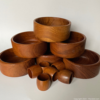 Photo shows six round teak wood bowls stacked in a pyramid shape with six smaller teak napkin rings piled in front. Rich brown wood grain visible.