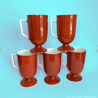 Five burnt orange pedestal base mugs with white handles arranged together on a blue background.