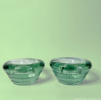 Pair of emerald green glass tea light candle holders shown side by side on light green background. Each is round and thick glass with clear green color and recessed top for tea light candle.