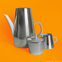 Front view of stainless steel and porcelain tea set components showing reflective metallic and cream/white finishes against vibrant orange background.