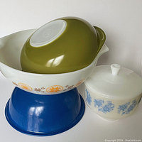 Four Pyrex kitchenware items are displayed: three mixing bowls (white with orange/yellow floral pattern, blue solid color, olive green solid color) and one lidded casserole dish with blue floral pattern on white ceramic.