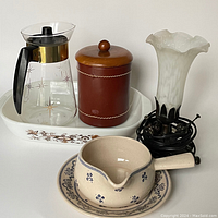 Photo showing a glass coffee carafe, wooden canister, ceramic saucepan with saucer, and a decorative vase on a white tray.