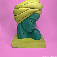 Side view of the chalkware bust lamp base showing yellow turban, green face and shoulder detail, and square base on pink background.