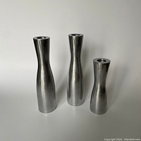 Photo showing three stainless steel candlestick holders of varying heights with smooth, polished surfaces on a white background.