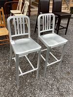Pair of silver aluminum bar height chairs displayed outside among other assorted chairs. Shows front and side views of each chair to highlight shape and structure.