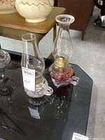 Photo showing the pair of vintage glass finger lamps on a black glass table with their clear glass chimneys, one lamp with red glass base and the other clear pressed glass base.