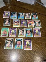 Photo of 18 vintage Garbage Pail Kids collectible cards laid out on a wood surface showing full designs and colors.