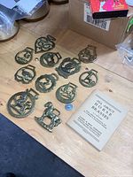 Top view of 11 brass horse brasses spread on a wooden table with a book 'All About Horse Brasses' nearby.