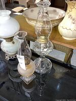 Photo showing smaller hobnail patterned oil lamp and larger clear glass oil lamp with chimneys on reflective surface with floral ceramics in background.