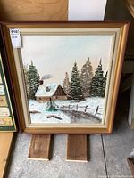 Full view of the framed oil painting depicting a winter scene with a cabin, snow, trees, and a fence.