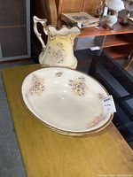 Photo showing antique porcelain pitcher and oval wash basin on a table, both decorated with floral patterns and gold trim.