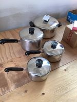 Four stainless steel pots with lids, black handles, and copper bottoms arranged on a wooden table.