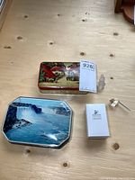 Two vintage tins on wood surface; waterfall scene 'Ruby Toffee' tin and home scene tin, with Swarovski box and a small crystal bell outside.