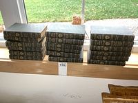 Eighteen black hardcover books arranged in three stacks by a window. Each book spine has gold titles and Harvard University crest.