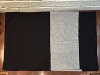 Full view of the rectangular shawl laid flat with black on left and right sides and grey panel in center, on wooden table beneath white textured wall.