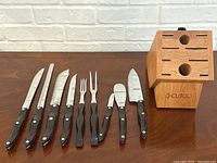All nine Cutco utensils laid out next to oak block on table against white brick background