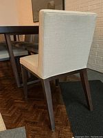 Rear three-quarter view of one chair showing the cream woven upholstery and dark stained wooden legs next to a dining table