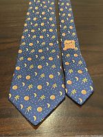 Full view of Hermes blue silk tie with orange fruit, moon, and palm tree pattern and visible Hermes Paris label.