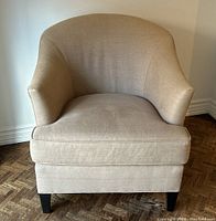Front view of beige upholstered barrel-back chair with removable cushion and black wooden legs