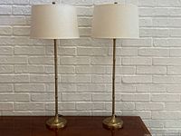 Front view of both brass table lamps with off-white drum shades against white brick wall